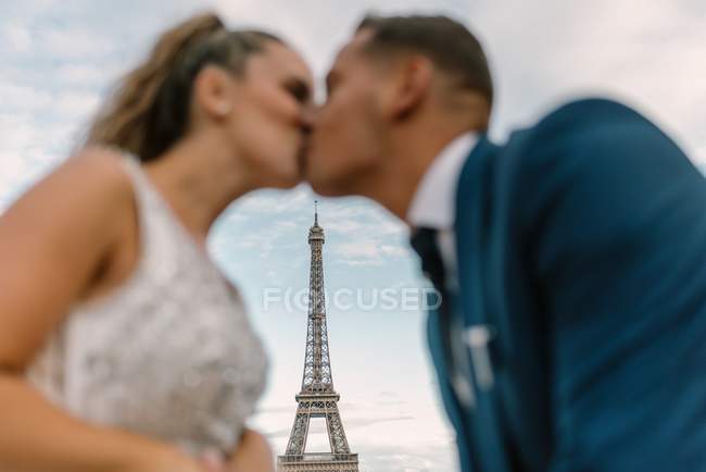 Out of focus groom in blue suit and bride in white wedding gown kissing passionately with Eiffel Tower on background at Paris — Stock Photo