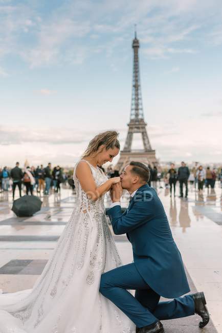 Contenuto sposo in abito elegante blu in piedi sul ginocchio e baciare le mani della sposa soddisfatta in abito da sposa bianco con Torre Eiffel sullo sfondo — Foto stock