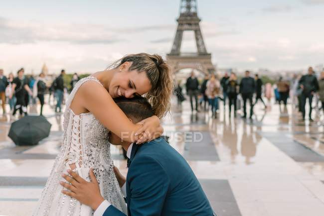 Contenu marié en costume élégant bleu debout sur le genou et embrasser mariée satisfaite en robe de mariée blanche avec Tour Eiffel sur fond — Photo de stock