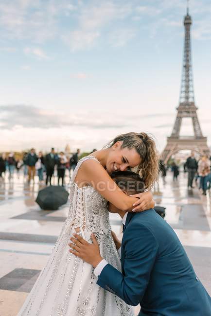 Contenu marié en costume élégant bleu debout sur le genou et embrasser mariée satisfaite en robe de mariée blanche avec Tour Eiffel sur fond — Photo de stock