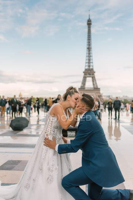 Novio en traje azul arrodillado y novia en vestido de novia blanco besándose apasionadamente con la Torre Eiffel en el fondo en París - foto de stock