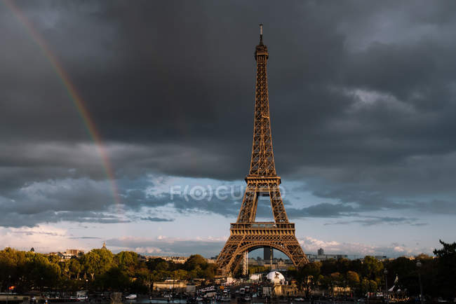 Powerful Eiffel Tower among green plants and buildings with cloudy sky and rainbow on background at Paris — Stock Photo