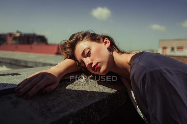 Modelo femenina joven sin emociones con labios rojos en ropa casual apoyada en una valla balanceada con cielo azul sobre fondo borroso en el techo de los edificios - foto de stock