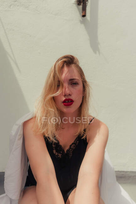 Fashionable female blonde haired model with beautiful red lops in stylish black tank top sitting and looking at camera with white wall on background — Stock Photo