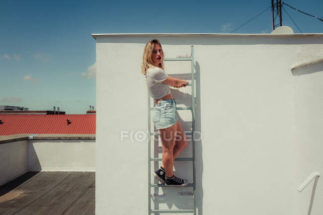 Joven adolescente en pantalones vaqueros casual camiseta blanca y zapatillas de deporte subir la escalera en el techo del edificio y mirando a la cámara en el día soleado de verano - foto de stock