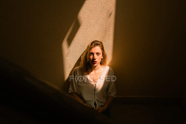 Sensibilità giovane donna con labbra rosse in giacca bianca casual seduta in camera buia con bagliore di luce sul viso sfondando la finestra e guardando la fotocamera — Foto stock