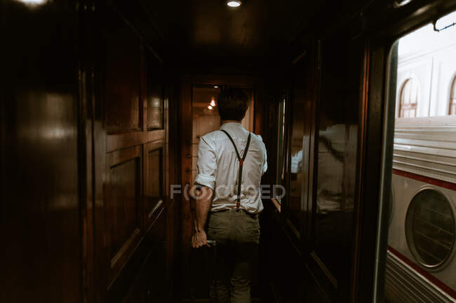 Visão traseira do homem anônimo em roupas vintage puxando bagagem e andando no corredor do trem antigo — Fotografia de Stock