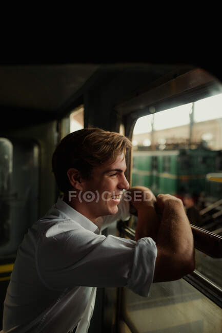 Vista lateral de hombre joven feliz en camisa sonriendo y mirando por la ventana del tren durante la parada en la estación de tren - foto de stock