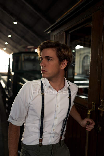 Young man in retro outfit looking away before entering aged train on platform of railway station — Stock Photo