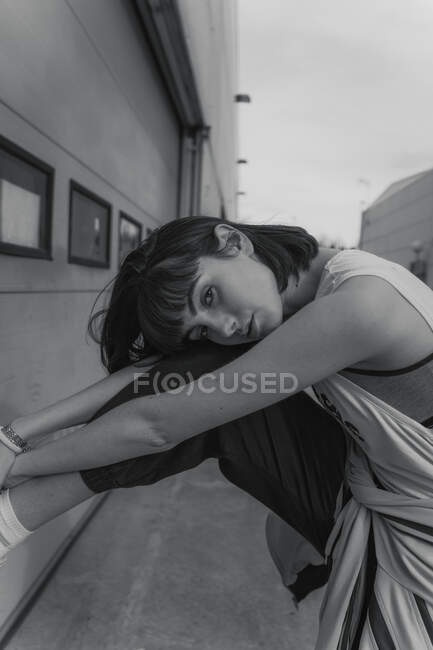 Черно-белый вид на элегантную женщину-подростка в повседневной одежде, опирающуюся на стену здания и опирающуюся на руки и голову на ногу, глядя в камеру на городской улице — стоковое фото
