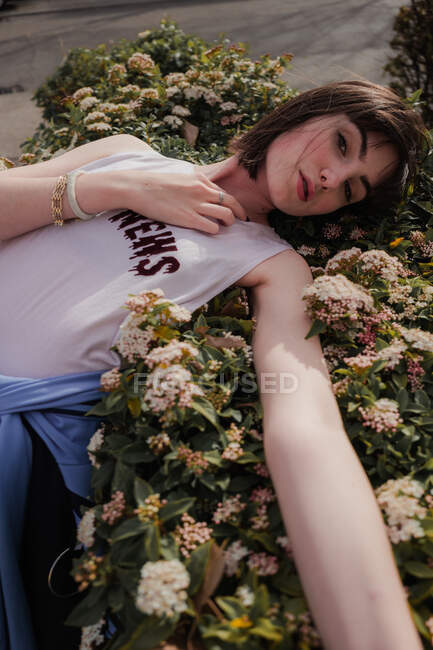 Зверху елегантна жінка-підліток в повсякденному стильному одязі, спираючись на квітучий зелений кущ і дивлячись на камеру на міській вулиці — стокове фото