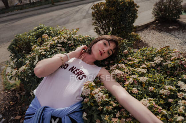 Сверху элегантная девушка-подросток в повседневной стильной одежде опирается на цветущий зеленый кустарник и смотрит в камеру на городской улице — стоковое фото