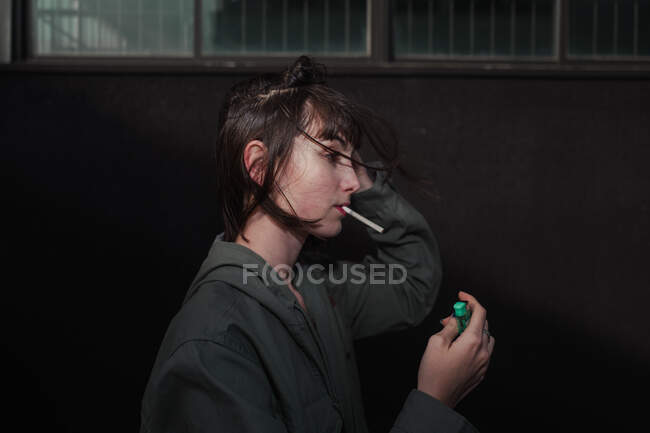 Vista lateral da jovem morena rebelde na elegante jaqueta casual com pregos azuis acendendo cigarro na rua da cidade — Fotografia de Stock