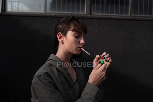 Side view of rebel young brunette in stylish casual jacket with blue nails lighting up cigarette at city street — Stock Photo