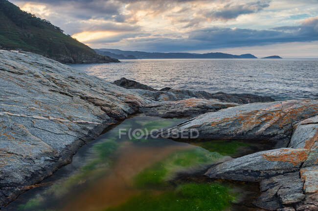 From above of small pound with moss among powerful rocks with calm sea overgrown mountains and cloudy sky on background at Portonovo beach — Stock Photo