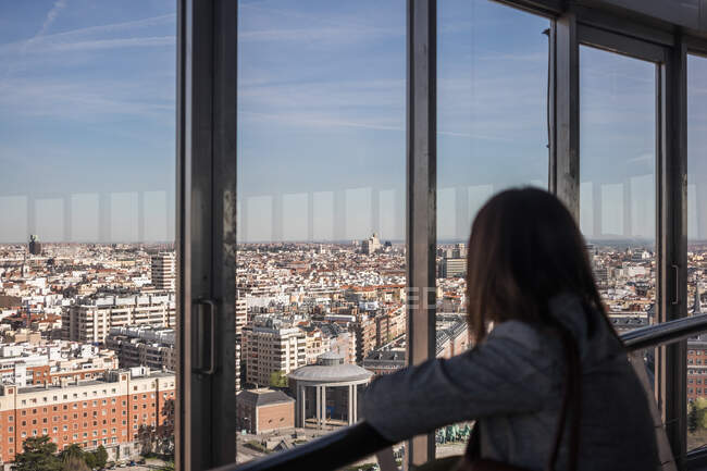 Mulher irreconhecível apoiando-se em trilhos e admirando a cidade moderna através da janela da varanda no dia ensolarado — Fotografia de Stock