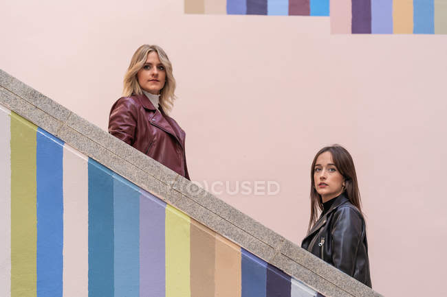 Side view of attractive pensive young friends in jackets standing on different level of striped colored stairs outdoors looking at camera — Stock Photo