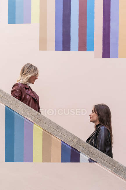 Вид сбоку привлекательных задумчивых молодых друзей в куртках, стоящих на разных уровнях полосатых цветных лестниц на открытом воздухе, смотрящих друг на друга — стоковое фото
