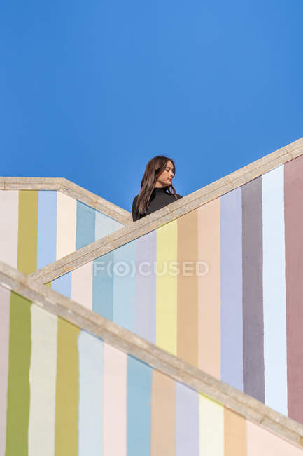 Vue latérale de la jeune femme attrayante et coûteuse en vestes debout sur différents niveaux d'escaliers colorés rayés à l'extérieur regardant loin — Photo de stock