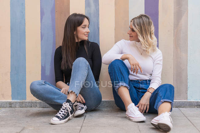 Sonriendo mujeres elegantes positivas apoyadas una en la otra mientras se sientan cerca de la acera cerca de la pared de la calle rayada mirándose entre sí - foto de stock