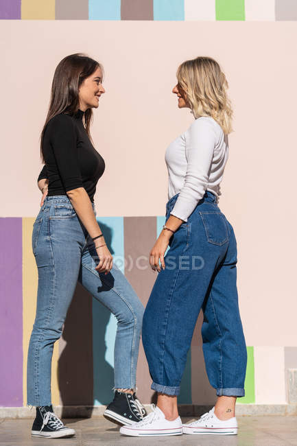 Smiling positive elegant women standing while leaning on stripped colorful wall close to sidewalk on the street looking at each other — Stock Photo