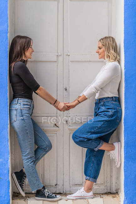 Side view of happy gentle caring girlfriends in stylish outfit holding hands together while standing near door looking at each other — Stock Photo