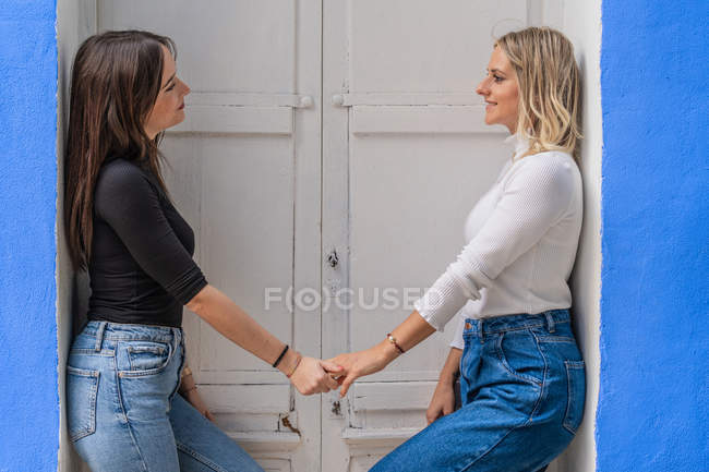 Seitenansicht von glücklichen sanft fürsorglichen Freundinnen in stylischem Outfit, die sich die Hände halten, während sie in der Nähe der Tür stehen und einander anschauen — Stockfoto