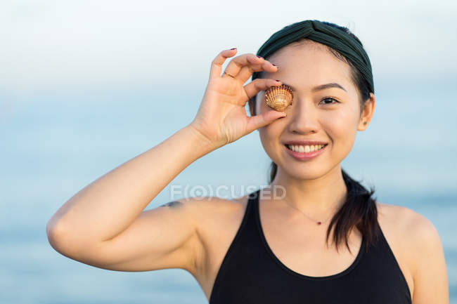 Азиатская женщина в спортивной рубашке смотрит в камеру и закрывает глаз морской ракушкой, стоя на пляже — стоковое фото