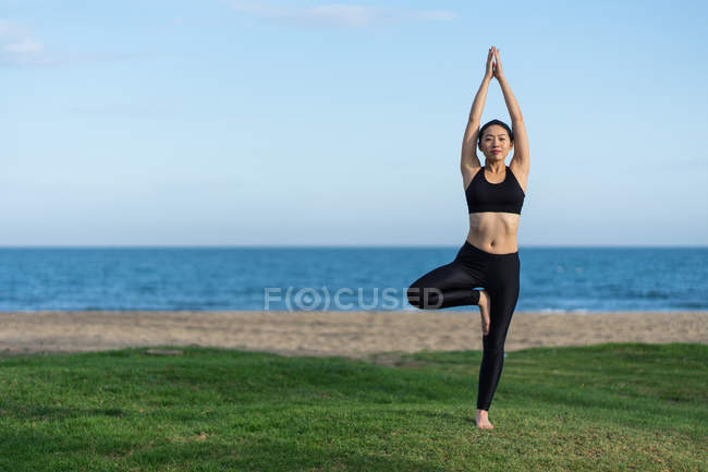 Giovane donna in top nero e leggings in piedi in posa albero su erba verde mentre pratica yoga sulla spiaggia — Foto stock