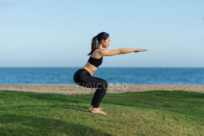 Young woman in black top and leggings standing on green grass practicing yoga on beach — Stock Photo