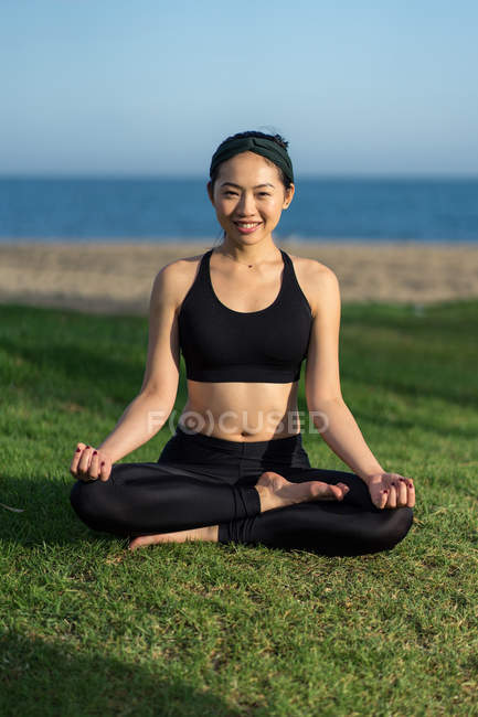 Mulher no topo preto e pernas sentadas na posição do lótus na grama verde com os olhos fechados enquanto medita na praia — Fotografia de Stock