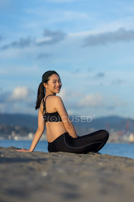 Vista laterale della donna in abbigliamento sportivo nero seduta con gambe incrociate sulla spiaggia riposante dopo l'allenamento guardando la fotocamera — Foto stock