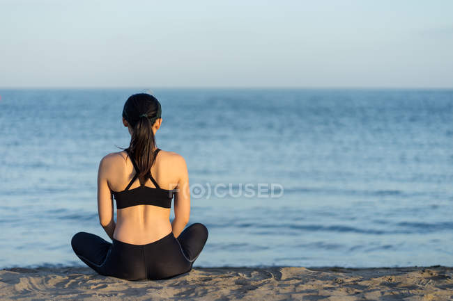 Rückenansicht einer Frau in schwarzer Sportbekleidung, die nach dem Training mit überkreuzten Beinen am Strand sitzt und sich ausruht — Stockfoto