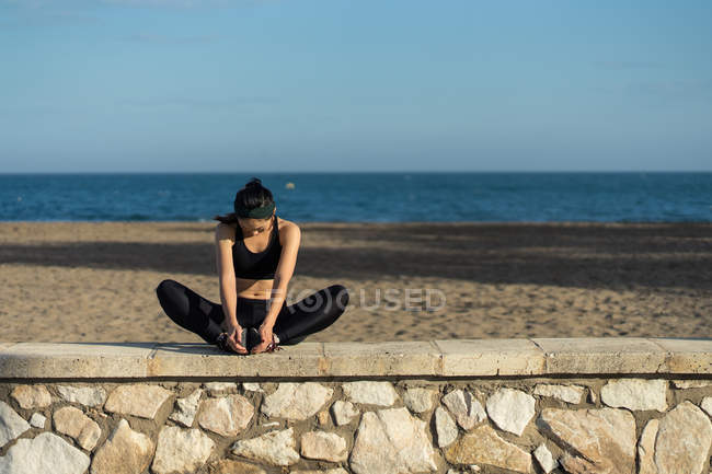 Jovem asiático mulher no preto topo e leggings fazendo alongamento exercício enquanto sentado ao lado de cerca de pedra à beira-mar — Fotografia de Stock