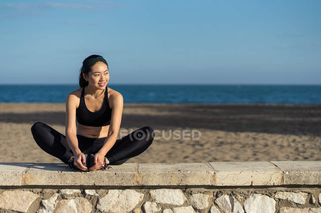 Mujer joven asiática en la cima negra y leggings que hacen ejercicio de estiramiento mientras se sientan junto a la valla de piedra en la playa. - foto de stock