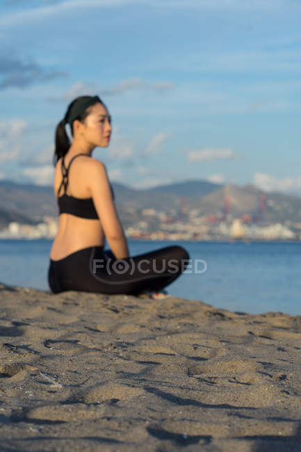 Вид сбоку на красивую женщину в черном платье, сидящую со скрещенными ногами на пляже, отдыхающую после тренировки — стоковое фото