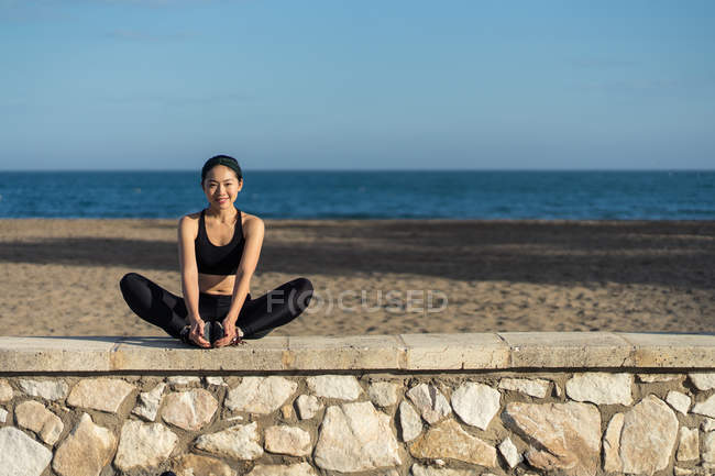 Молода азіатка на чорній верхівці і зябра роблять розтягування вправ, сидячи біля кам'яного паркану на узбережжі. — стокове фото