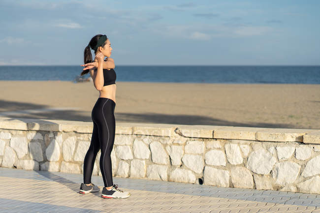 Vista lateral de la mujer asiática en la cima negra y las leyendas que hacen ejercicio de estiramiento mientras se encuentran junto a la valla de piedra en la playa. - foto de stock