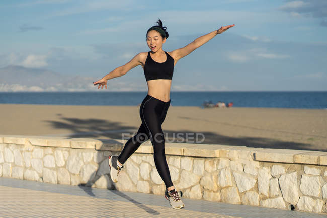 Gymnast in black leggings and crop top spreading hands to sides in jump on rocked path with fence — Stock Photo