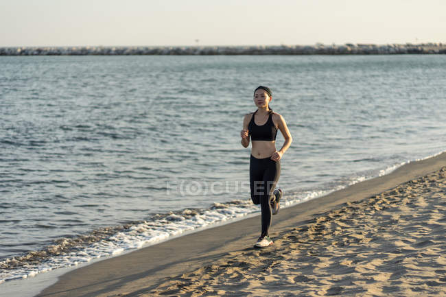 Motivated young female athlete in active black wear and sneakers jogging along sandy empty seashore — Stock Photo