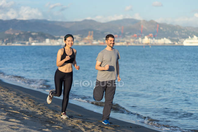 Couple in sports clothing smiling while jogging on sandy ocean side with city on opposite coast on blurred background — Stock Photo