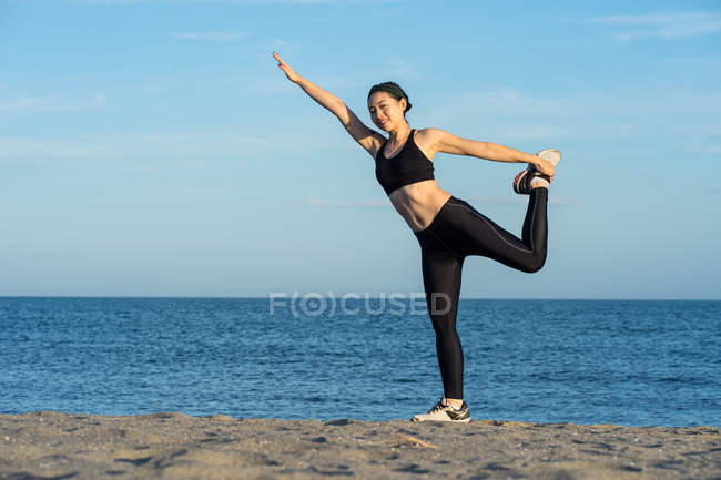 Strong young female athlete in black sports clothing standing on one leg while holding another leg with raised hand — Stock Photo
