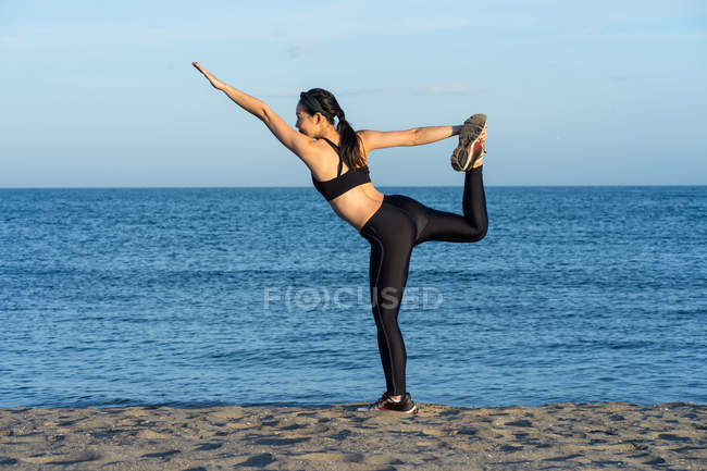 Jeune athlète féminine forte en vêtements de sport noirs debout sur une jambe tout en tenant une autre jambe la main levée — Photo de stock