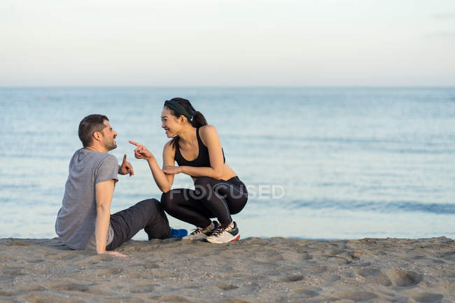 Весела молода багаторасова пара в спортивному одязі, сидячи на піщаному пляжі, відпочиваючи після тренування і насолоджуючись разом — стокове фото
