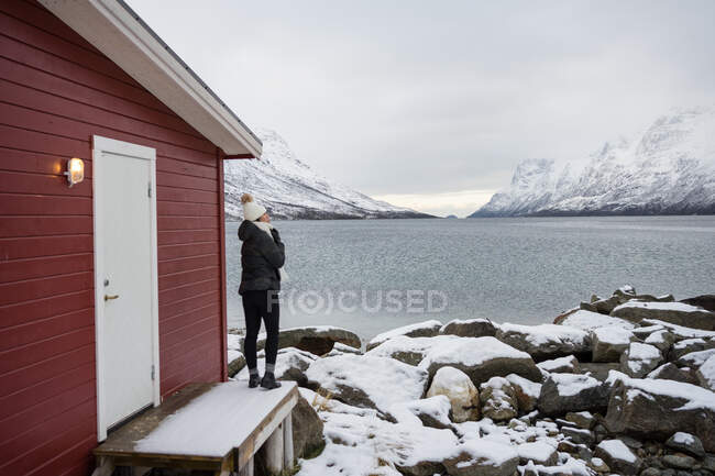 Mulher solitária na costa rochosa contra o lago tranquilo e montanhas nevadas no dia frio — Fotografia de Stock
