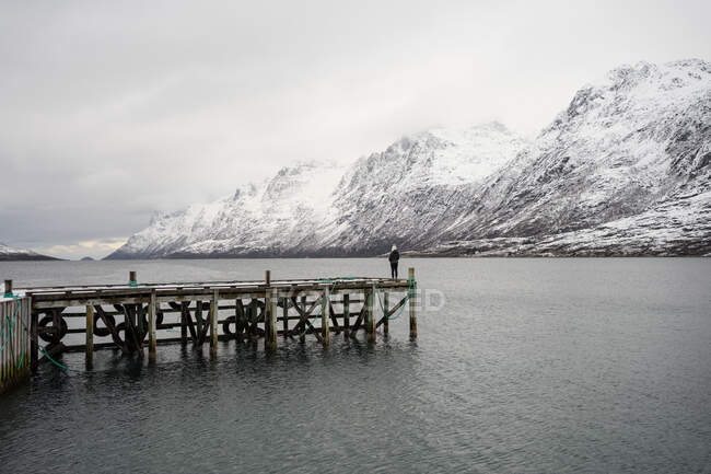 Самотній турист стоїть на дерев'яному пірсі посеред спокійного озера взимку — стокове фото