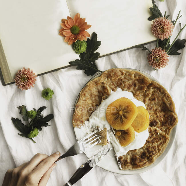 Top view of homemade crepes with cream and sliced persimmon served on plate with knife and fork on surface decorated with white cloth and flowers next to open book with empty pages — Stock Photo