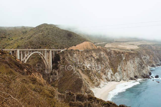 Blue waves washing rocky seashore with bridge in bright day — Stock Photo