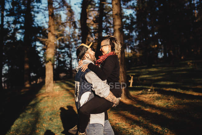Вид збоку бородатий чоловік тримає на руках веселу молоду леді і йде в ліс на розмитому тлі — стокове фото