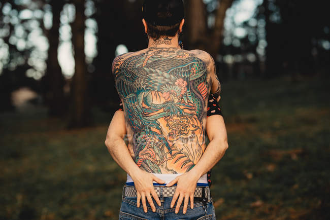 Vista posteriore del giovane uomo senza maglietta in tatuaggi con donna che abbraccia nel parco su sfondo sfocato — Foto stock